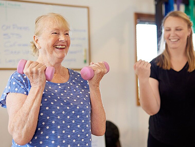 Elderly woman smiles while lifting weights. A younger woman demonstrates with weights beside her.