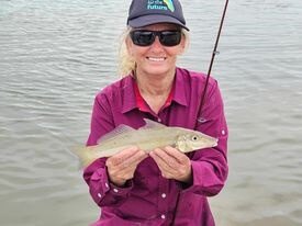 Woman dressed in purple long sleeve shirt and blue cap, smiles as she holds up a silver fish