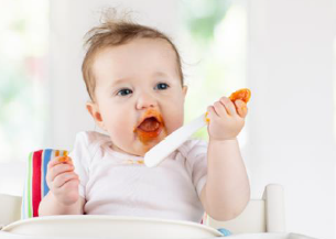 A baby sitting in a high chair holding a spoon eating pumpkin