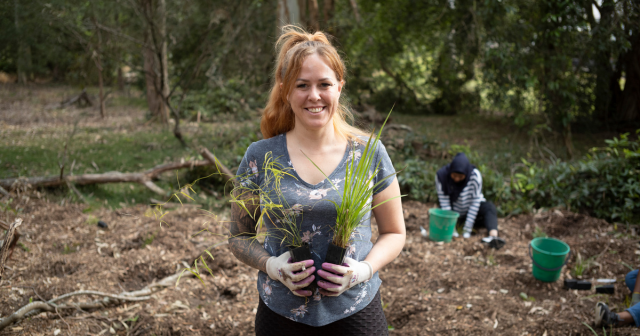 A woman in a green shirt happy and smiling holding plants in front of her with another women planting plants in the background