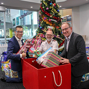 Three people are in front of a Christmas tree holding gifts