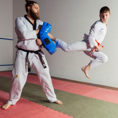 Child dressed in martial arts uniform jumps and kicks at a blue pad being held by an adult