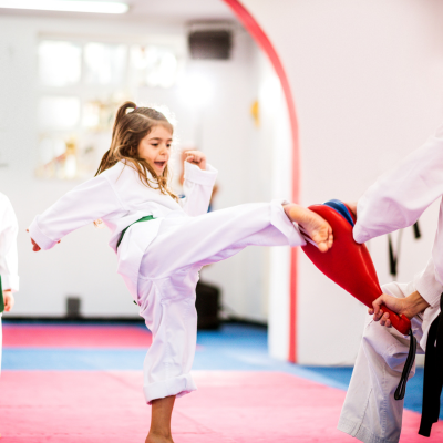 Girl dressed in white martial arts uniform kicks at another student holding kick pads