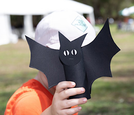 Child wearing an orange shirt and white hat holds up a black bat made out of black paper