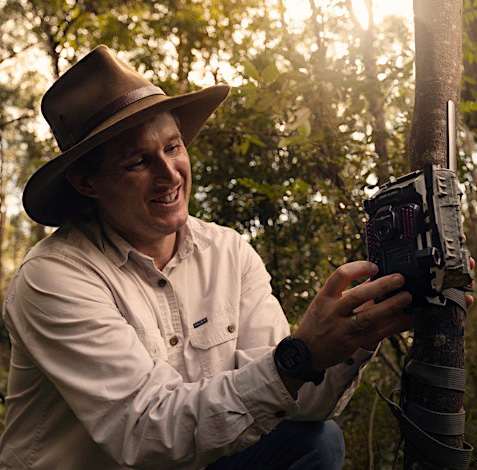 Smiling man dressed in a long sleeve khaki shirt and wide brimmed hat attaches a camera to a tree