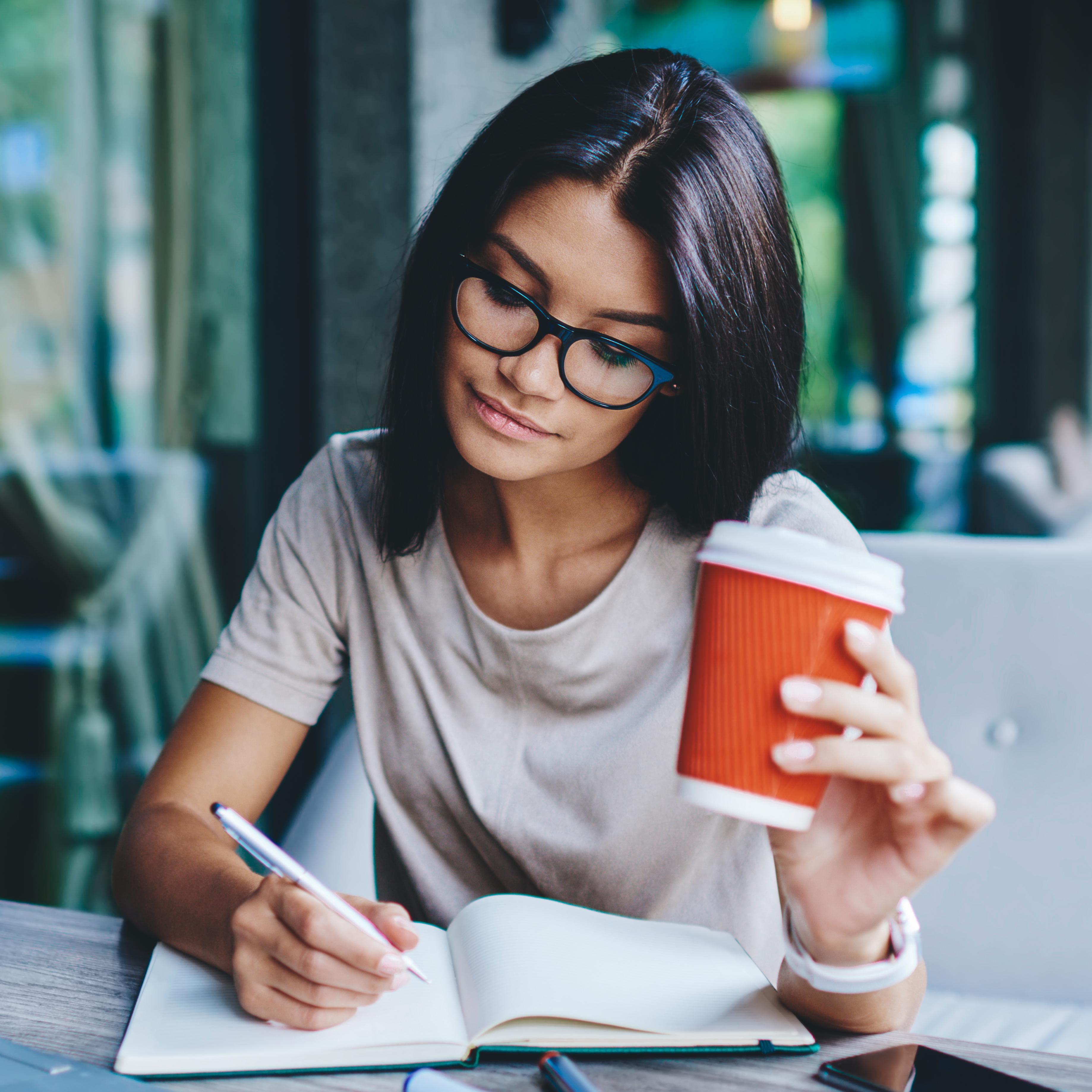 A girl writing in a book holding a coffee cup