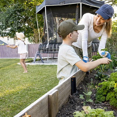 Woman and young child garden in raised garden beds while a young girl runs on the lawn beside a trampoline