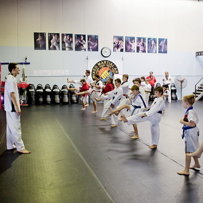 Young children lined up wearing white martial arts uniforms and attempting a kick while being watched by an instructor.