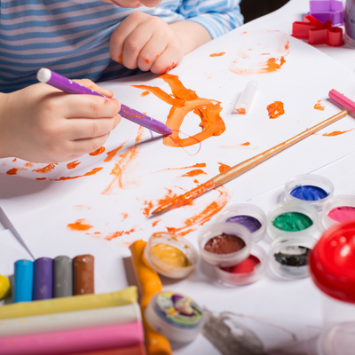Children's hands shown drawing on a sheet of paper with orange paint marks and other art supplies visible